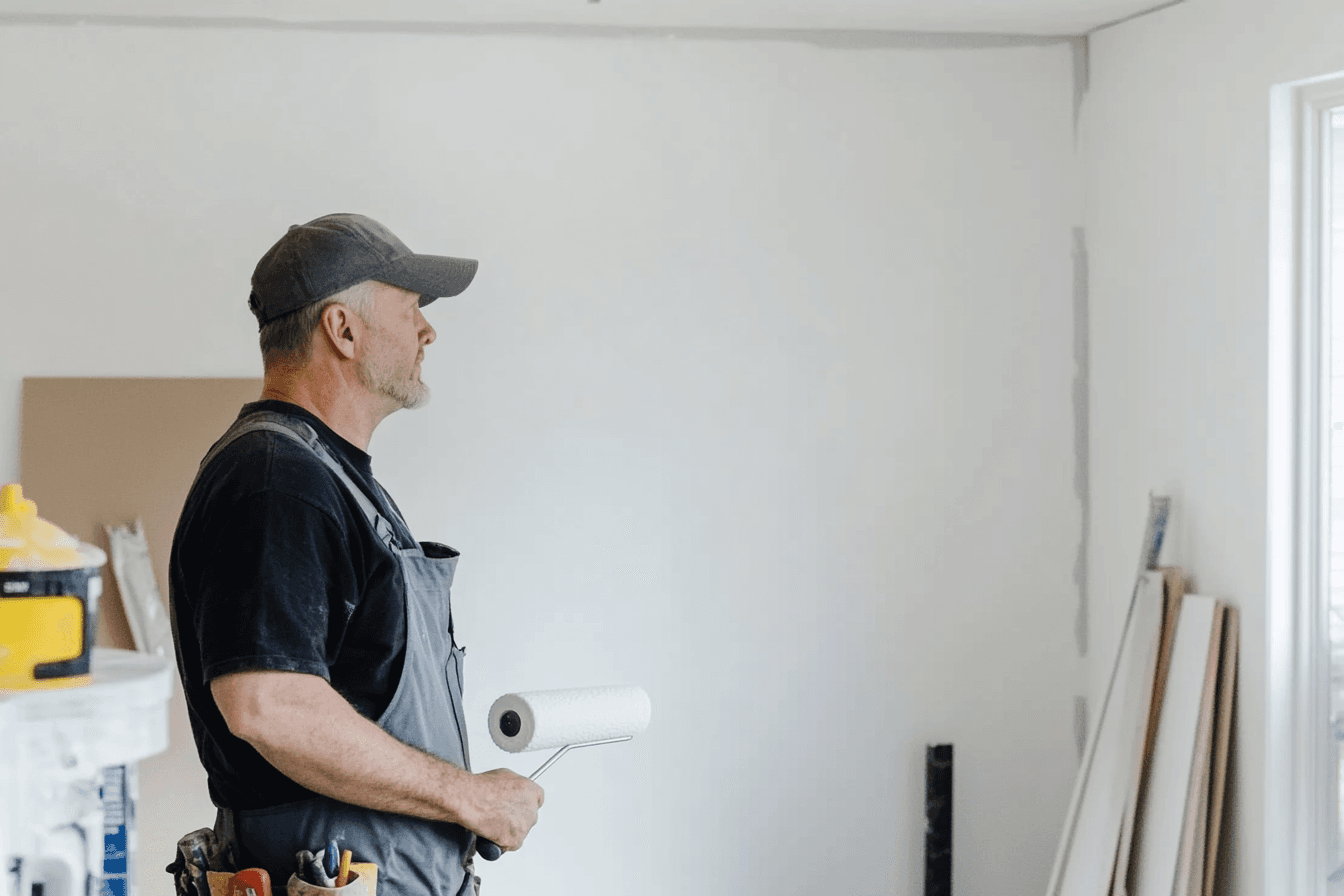 Peintre en bâtiment portant une salopette et une casquette, tenant un rouleau de peinture dans une pièce en rénovation.