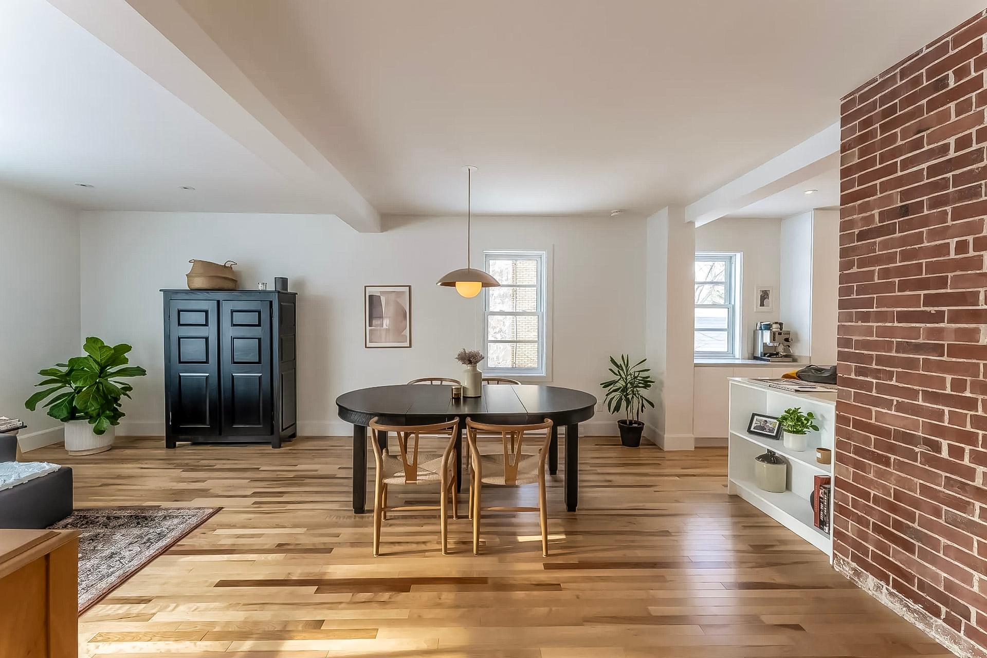Salle à manger moderne avec table noire, chaises en bois, parquet clair et mur en briques apparentes. Décoration minimaliste avec plantes d'intérieur et éclairage suspendu.