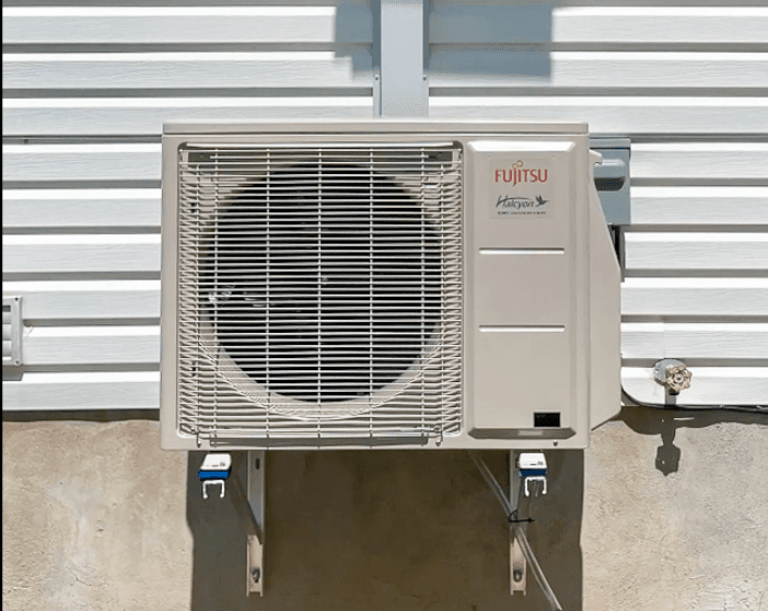 Unité extérieure de thermopompe Fujitsu Halcyon installée sur un mur blanc d'une maison résidentielle, montée sur un support métallique pour un système HVAC moderne, offrant chauffage et climatisation avec une efficacité énergétique optimale, assurant le confort thermique.